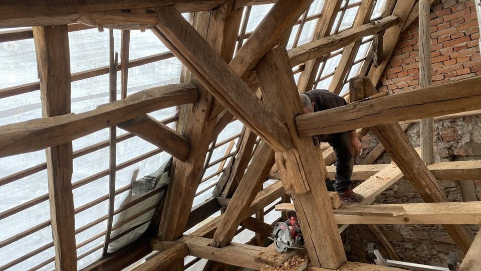 Blick in den historischen Eichendachstuhl: mehrere grob behauene Streben und Balken treffen an einem gezimmerten Knotenpunkt zusammen, dahinter arbeitet ein Zimmerer zwischen den Hölzern.